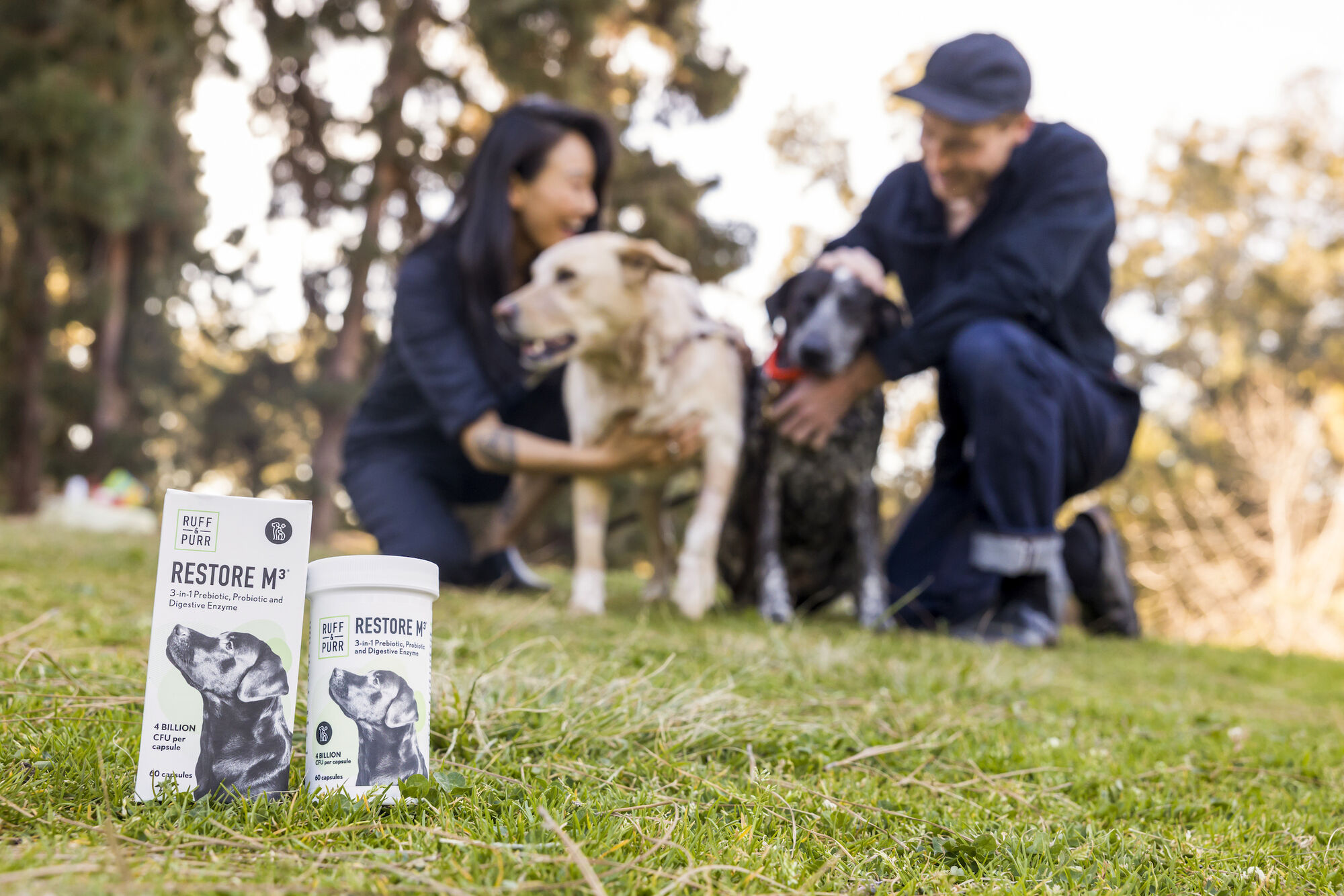 Photo of two humans playing with their two dogs with a bottle and box of Restore M3® by Ruff & Purr Pets in the foreground
