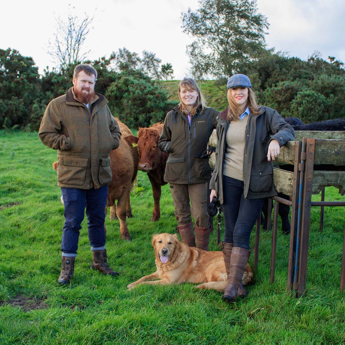 photo of a man and 2 womenin tweed jacket, wax jackets and leather boots, with cows standing behind and a dog in front.