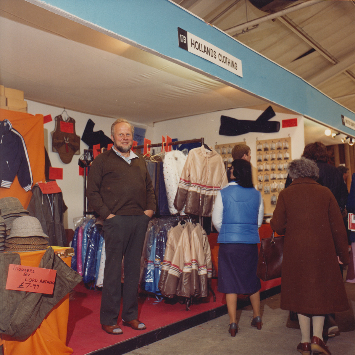 Man in brown jumper and trousers standing in front of a market stall named "Hollands Clothing".