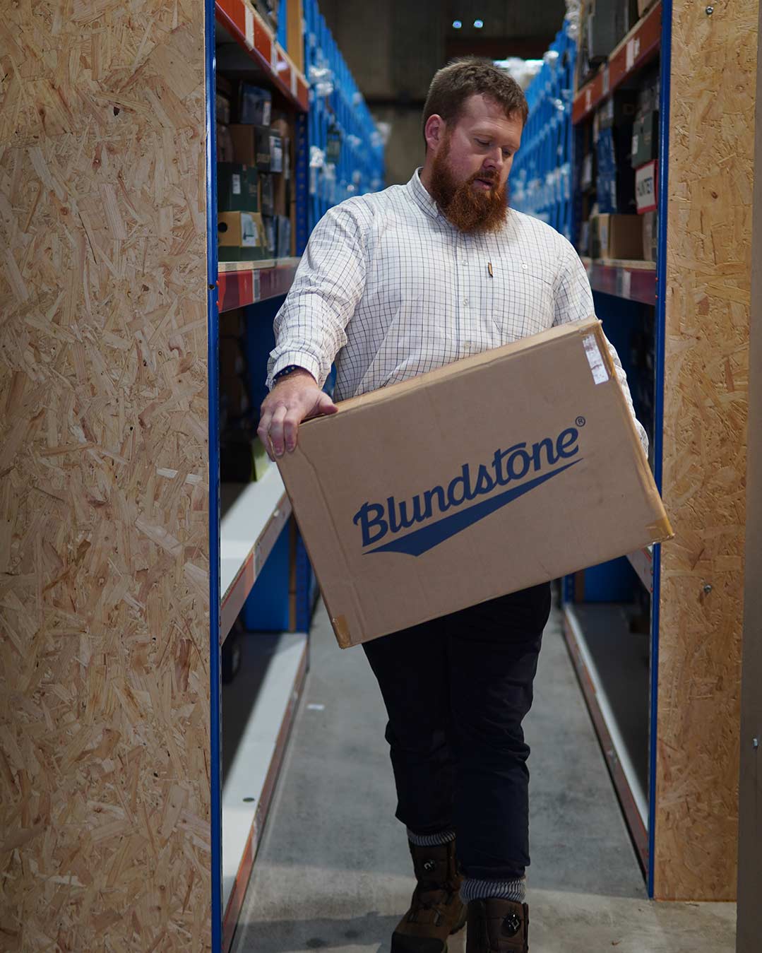 Matt Holland in tattersall shirt moving a box of Blundstone boots in a warehouse.